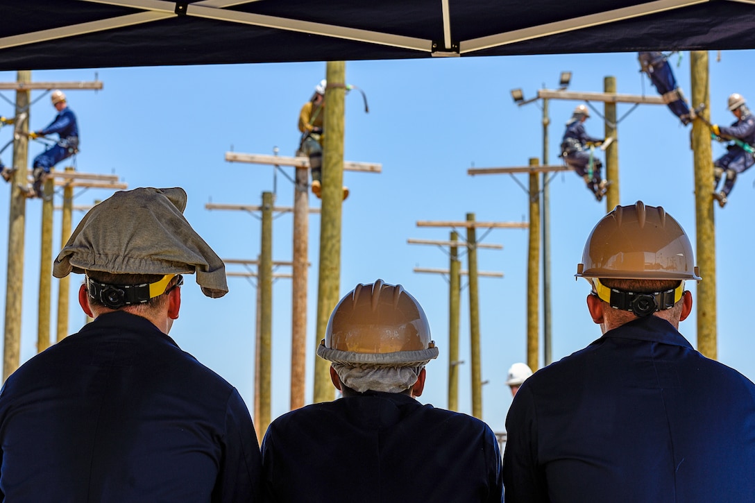 Three Navy recurits, shown from behind, watch fellow recruits climbing utility poles.