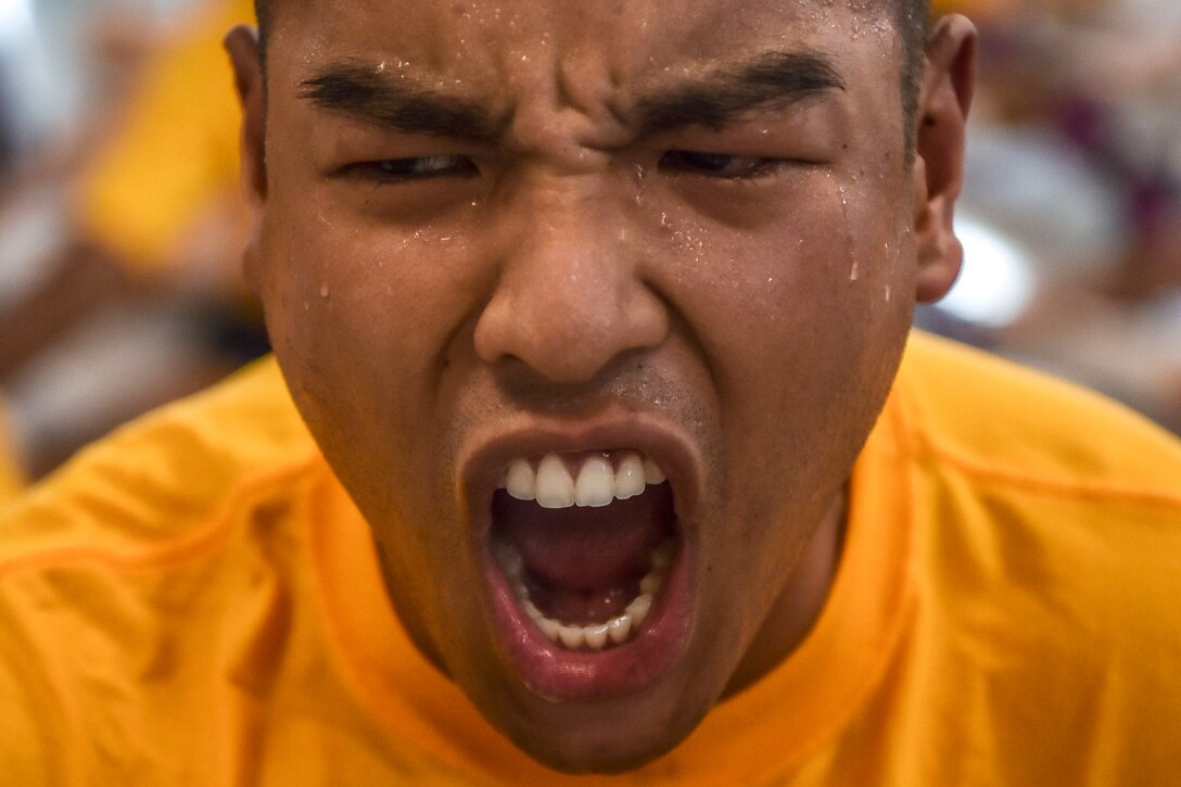 A Navy recruit, shown in closeup in a yellow t-shirt, yells.