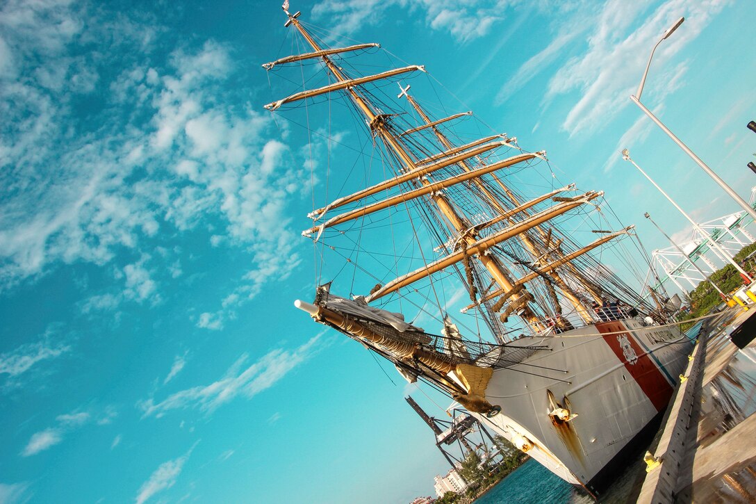 A tall ship, shown at an angle, sits moored at a pier, with bright blue sky in the background.