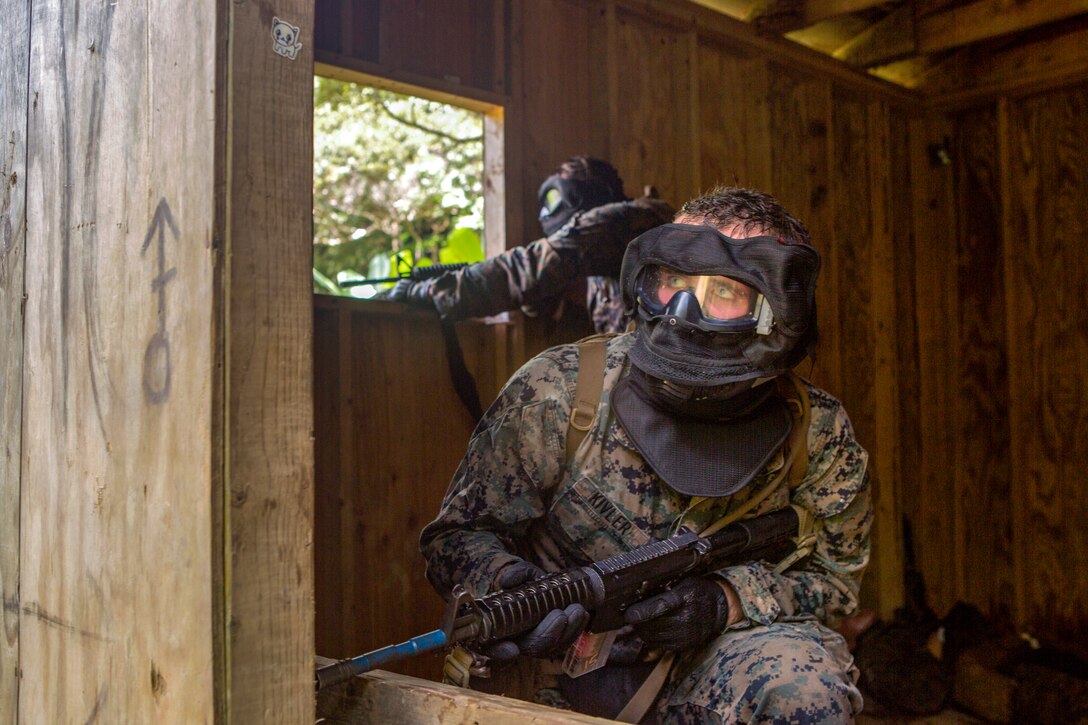 Marines in combat gear peek out from a wood structure.