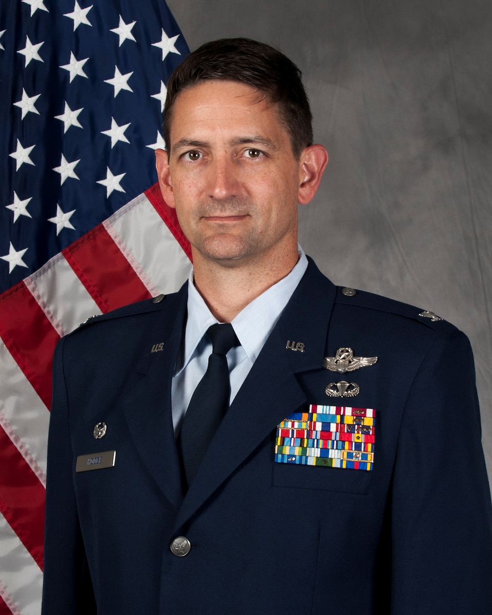 Dark haired white male Air Force colonel in blues in front of  red, white and blue American flag and grey background.