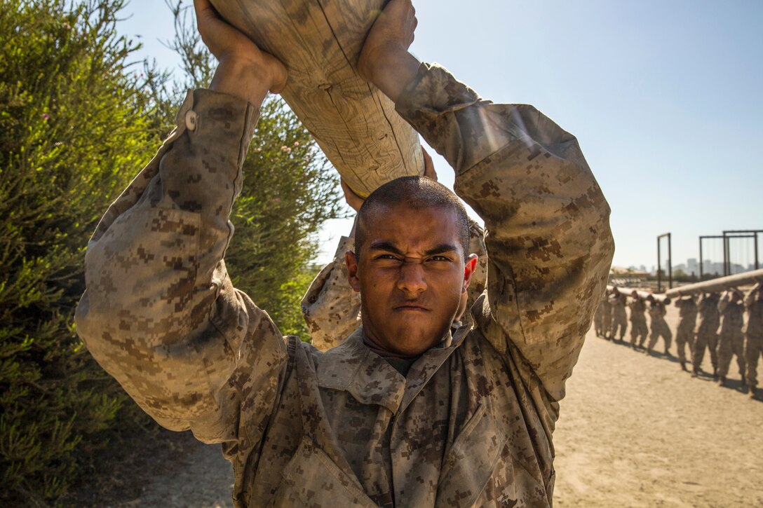 A Marine recruit grimaces while lifting a lot over his head with fellow recruits.