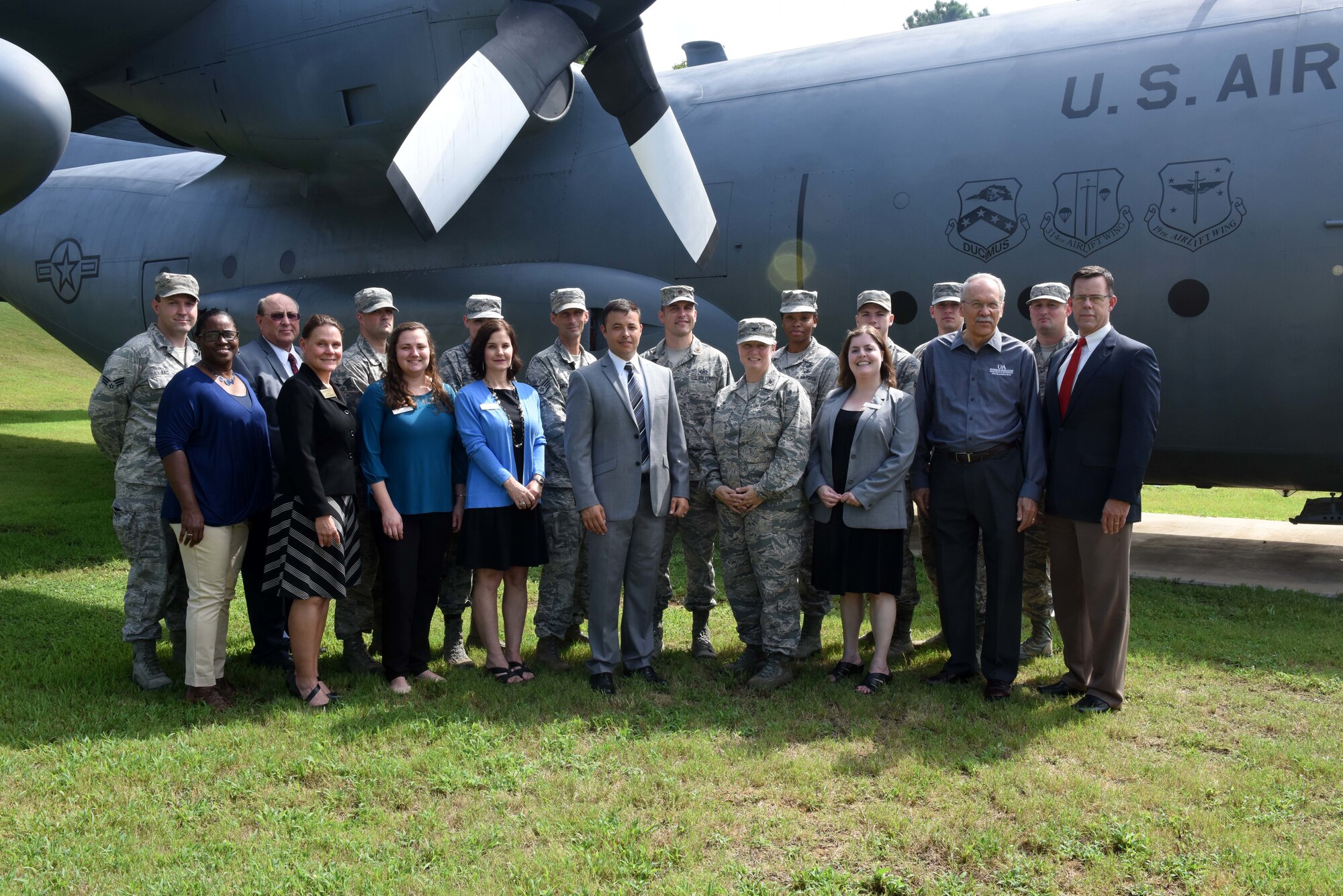 A group of individuals, both in uniform and in civilian business attire, stand in the grass in front of a large grey C-130H aircraft for a group photo.