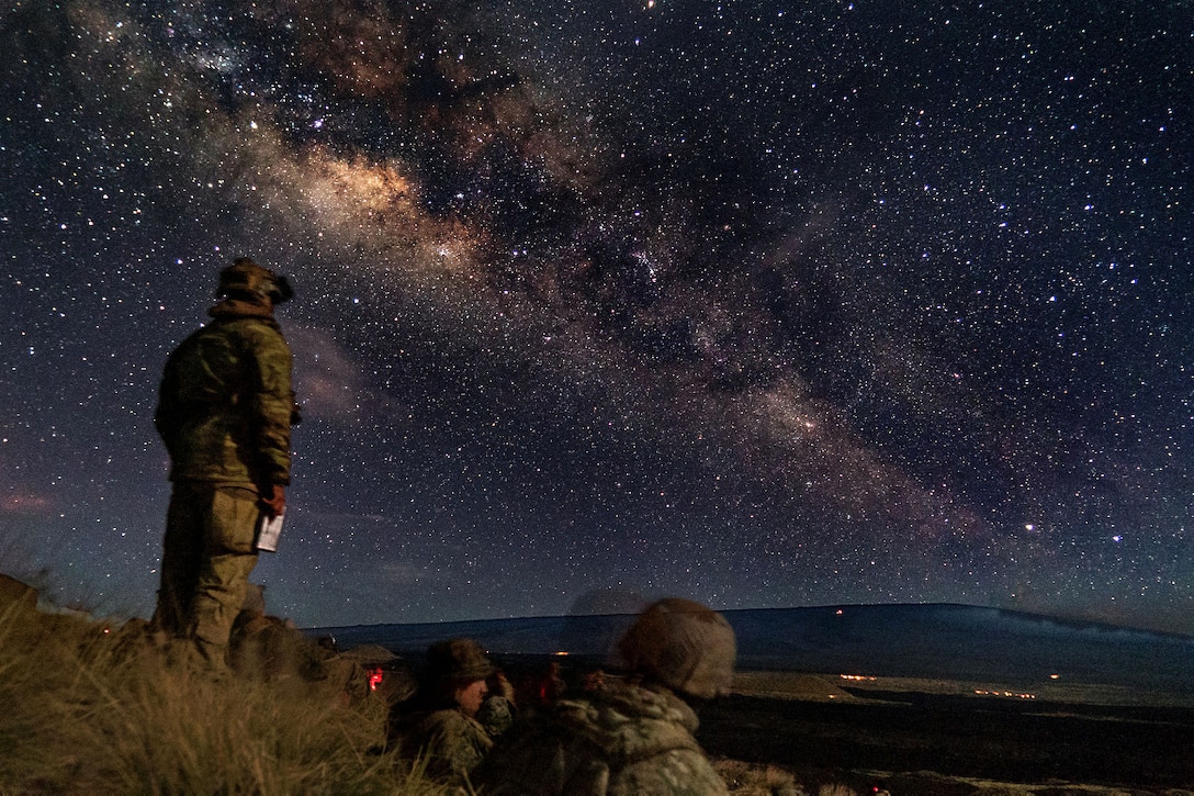 Service members sit and stand on a hill looking at stars and ordnance.