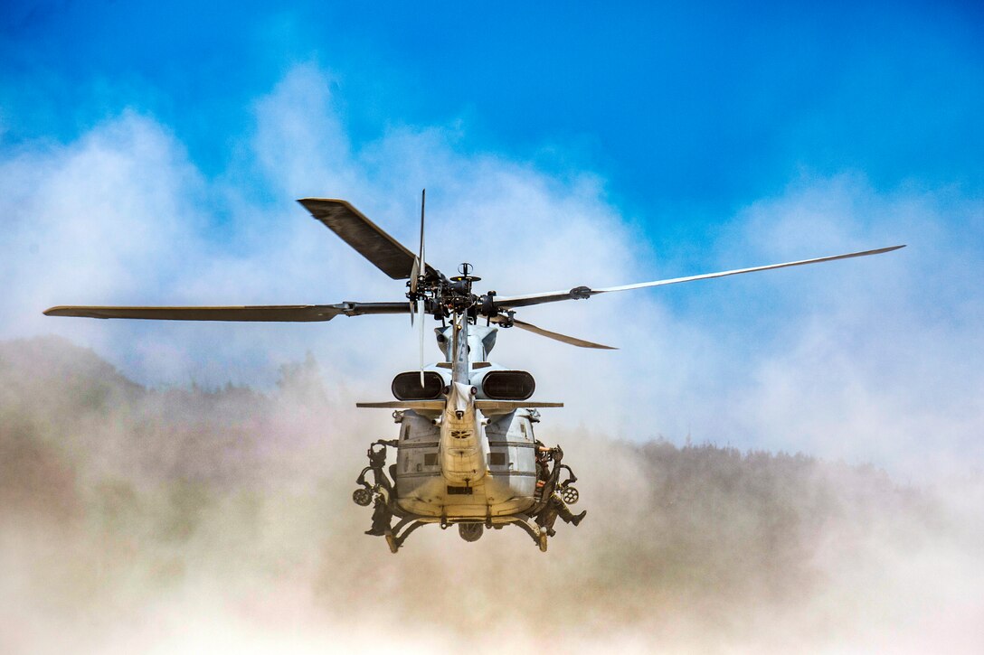 Marines hang out of a helicopter as it kicks up sand while taking off.