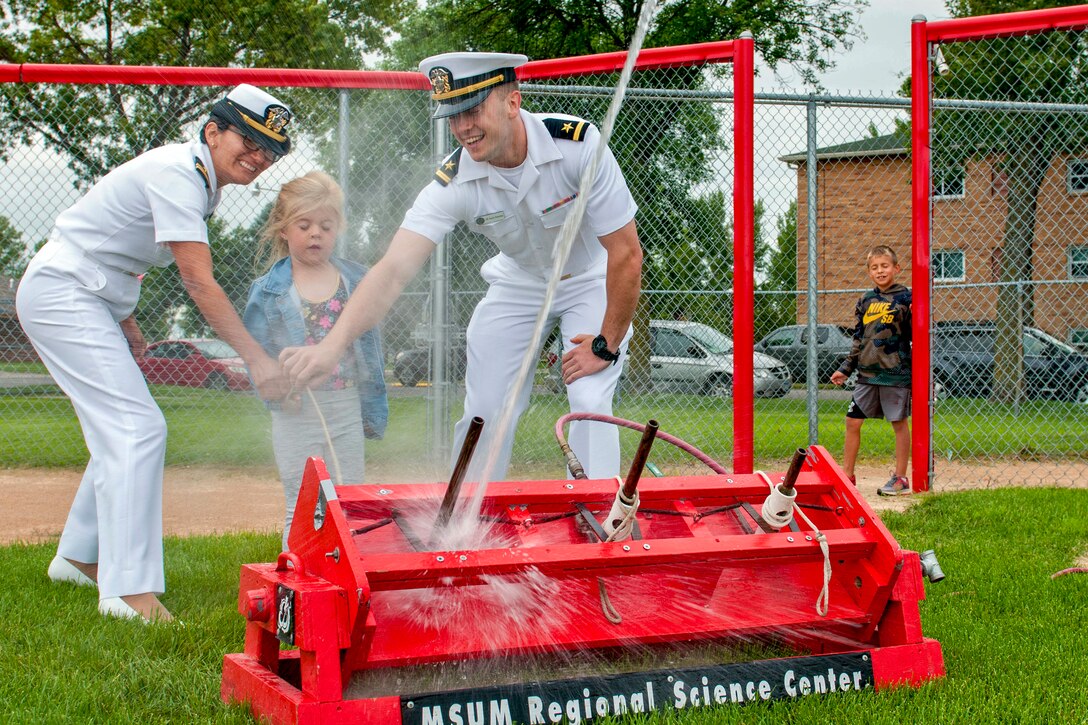 Two sailors and a child grasp a cord as a beam of water shoots from a red device on the ground in front of them.
