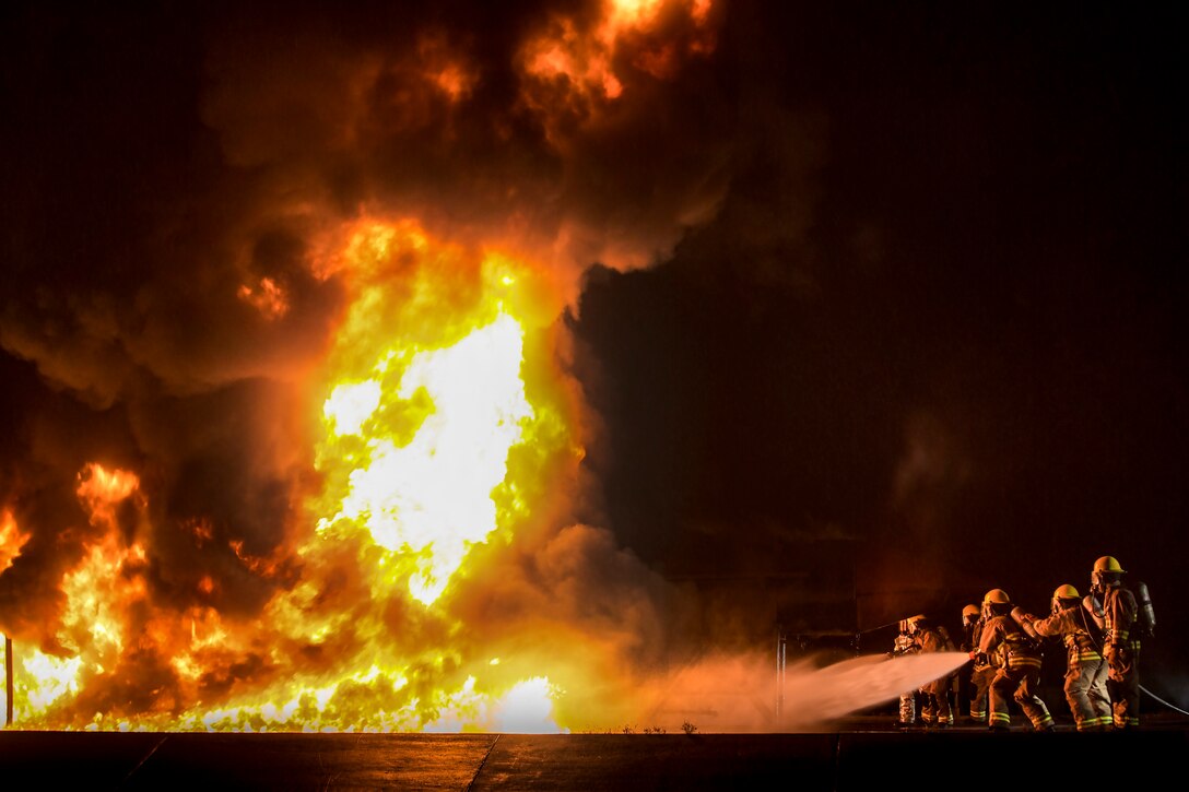 Marines wearing silver protective gear train a hose on a giant blaze.