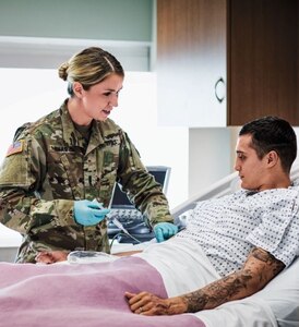 Caucasian female Officer Nurse 1LT Oskam standing bedside with male patient administering treatment inside a hospital.