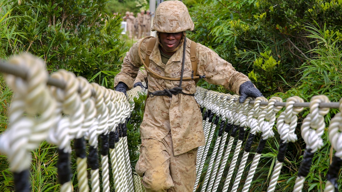 A Marine crosses a rope bridge amid green foliage.
