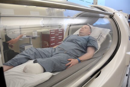 Air Force Staff Sgt. Maribel Cortez (right), certified hyperbaric technician, places the oxygen hood on Christen Hesser to demonstrate how a patient would receive oxygen therapy in the multi-person chamber at the new Undersea & Hyperbaric Medicine Clinic at Brooke Army Medical Center June 13, 2017. If the patient is not comfortable with the hood, they can also wear a mask to receive their treatment.