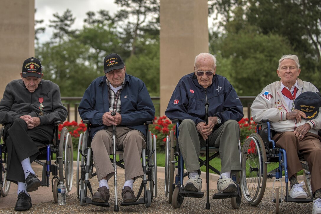 Four veterans sit in a row in wheelchairs at a memorial.
