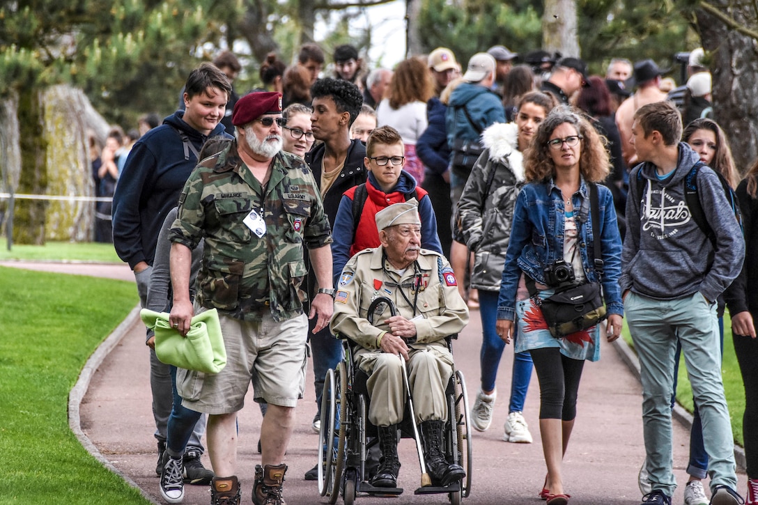Family members walk with a World War II veteran, who wears his uniform and sits in a wheelchair, on an outdoor path.