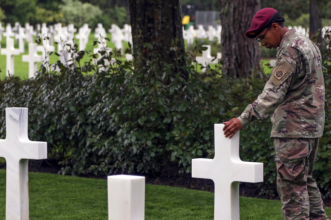 A soldier touches and looks down on a cross-shaped gravestone in a cemetery of identical stones.