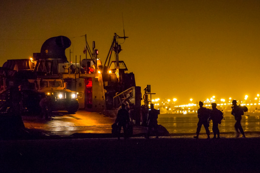 Troops, shown in silhouette, walk on shore near a vessel at night under a yellow sky illuminated by city lights in the distance.
