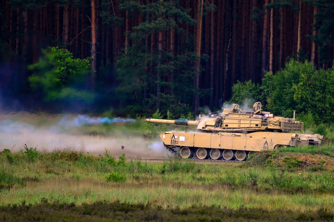 Smoke emanates from a tank's gun in a field in front of a forested area.