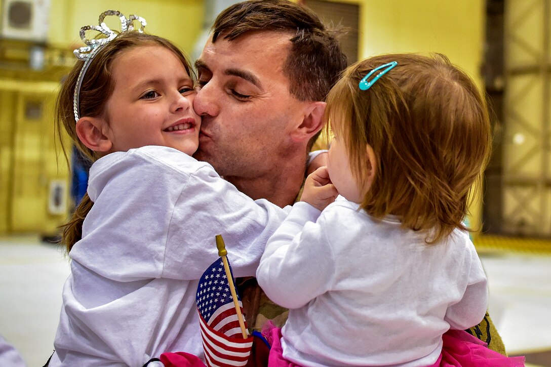 A soldier holds two little girls, kissing one of them on the cheek.