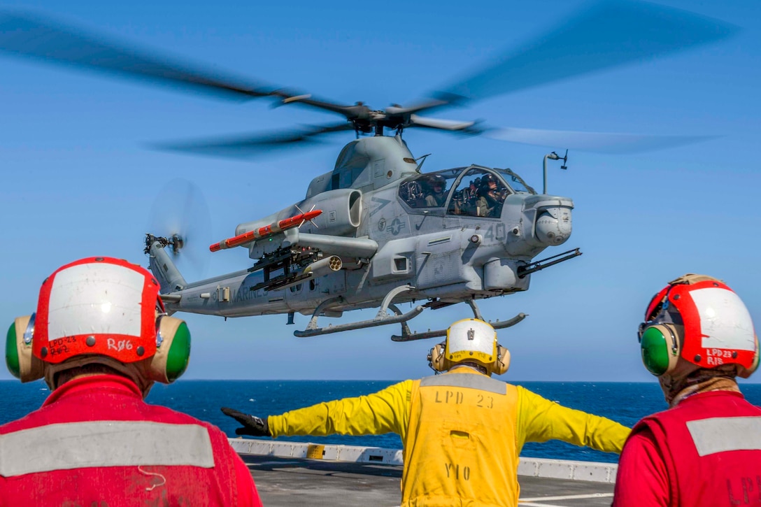 Three sailors stand on the deck of a ship while a helicopter lands.