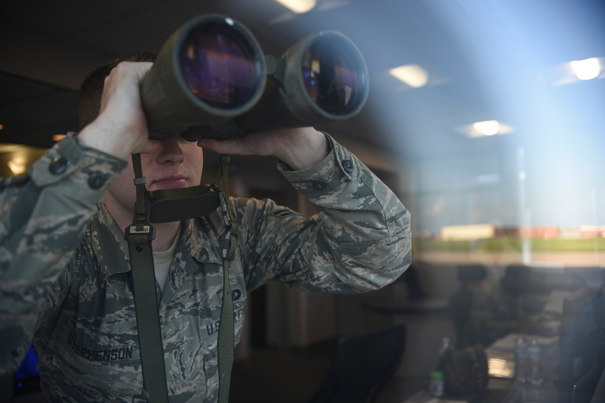 Airman 1st Class Casey Stephenson, 72nd Operations Support Squadron airfield management, uses a pair of binoculars to track airfield operations while part of the large hangars and runway he is watching is reflected in the glass May 23, 2018, Tinker Air Force Base, Oklahoma.