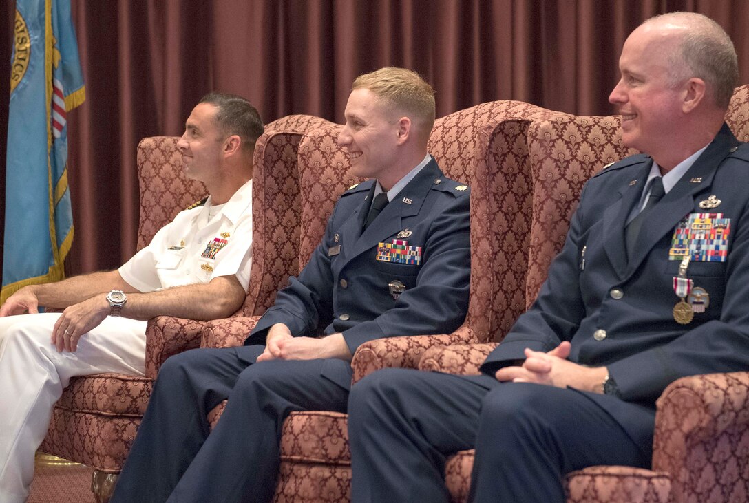 Three male military officers sit in high back chairs on a stage during a change of command ceremony.