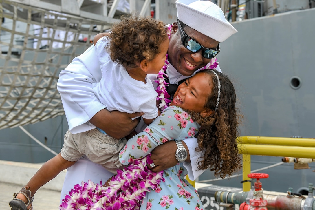A smiling sailor hugs a giggling girl and holds up a toddler by a ship on a pier.