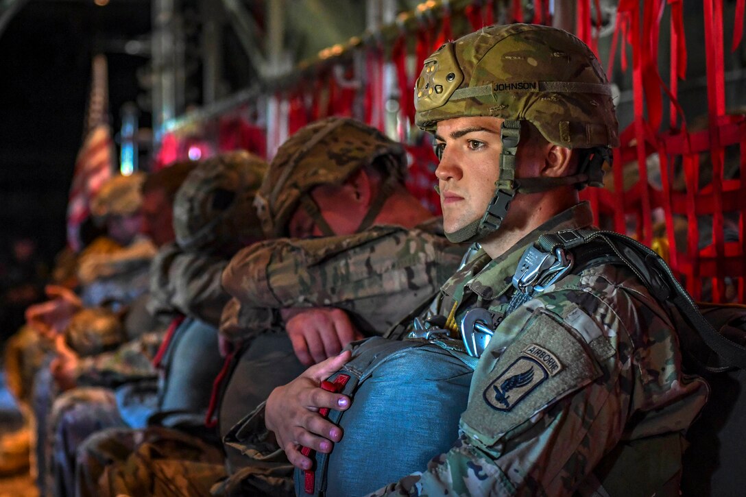 A soldier sits inside an aircraft with fellow troops and holds a parachute.