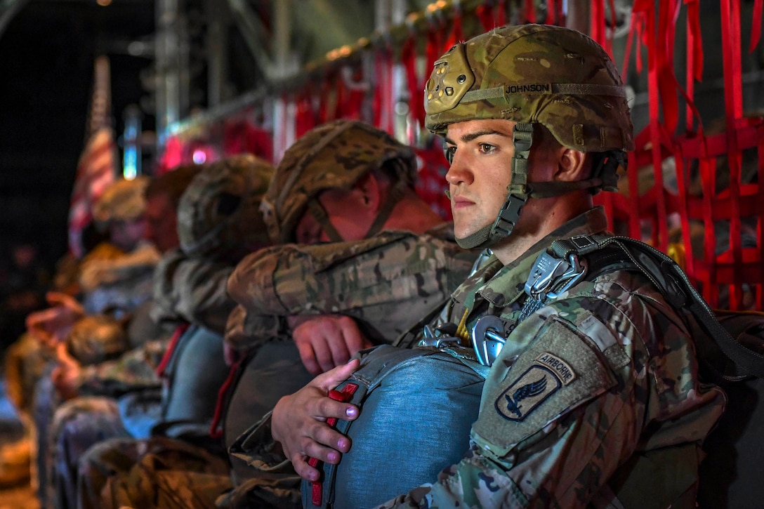 A soldier sits inside an aircraft with fellow troops and holds a parachute.