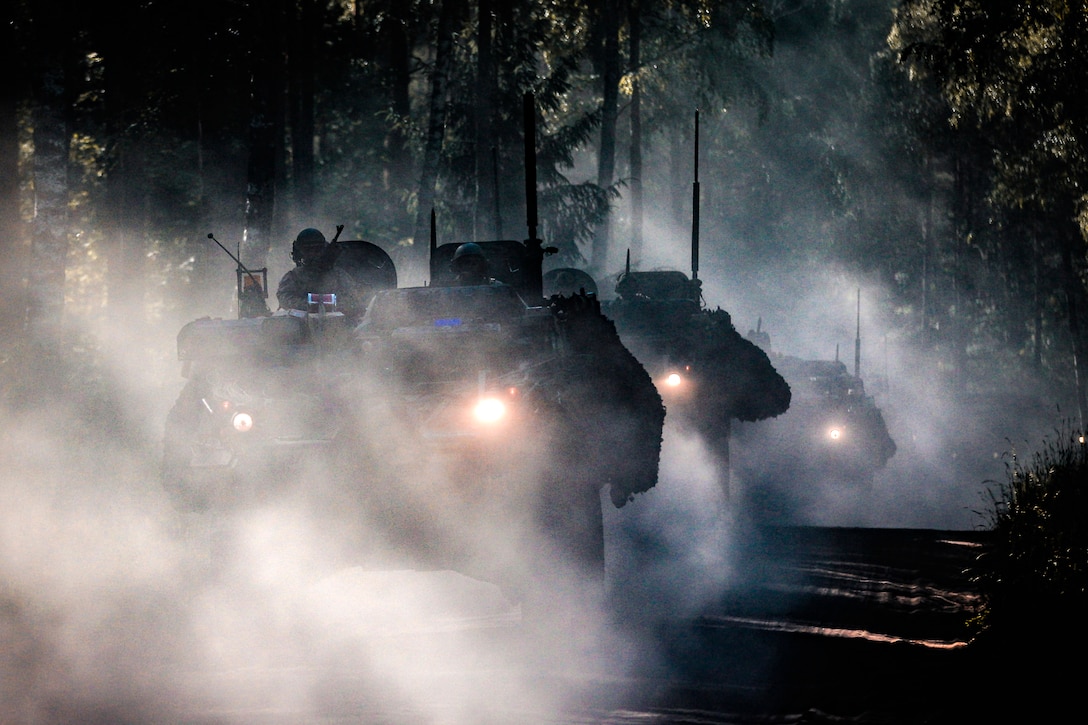 Military vehicles drive through thick fog on a road through forested area.