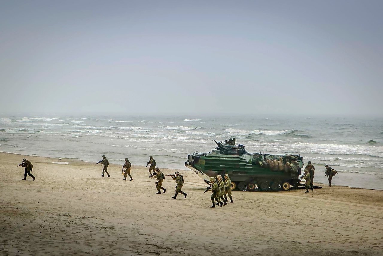 Troops point weapons while running on a beach away from a parked amphibious vehicle.