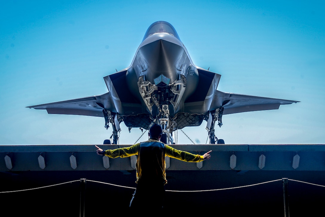 A sailor, shown from behind, holds his arms out as an aircraft sits on a raised platform in front of him.