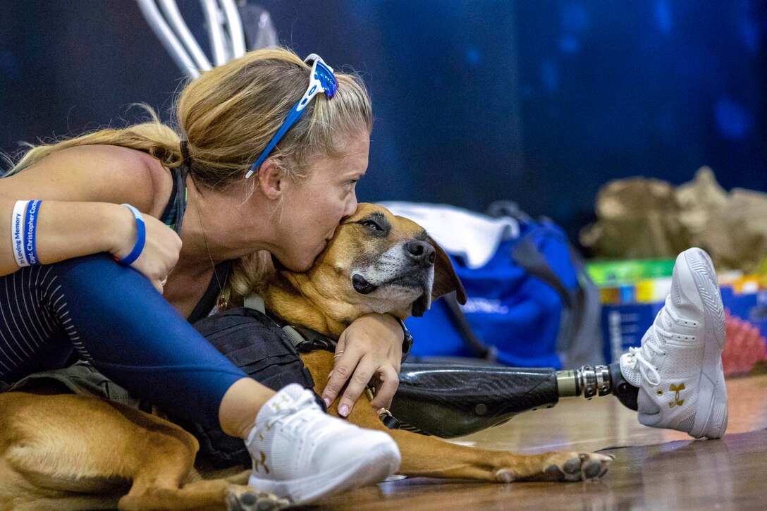 An airman sits on a gym floor and hugs and kisses a dog.