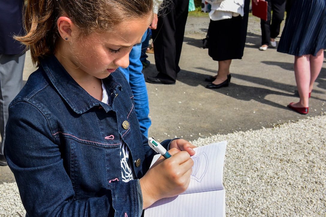 A local 8-year-old student draws a picture of Army Europe’s unit patch after the D-Day wreath-laying ceremony.