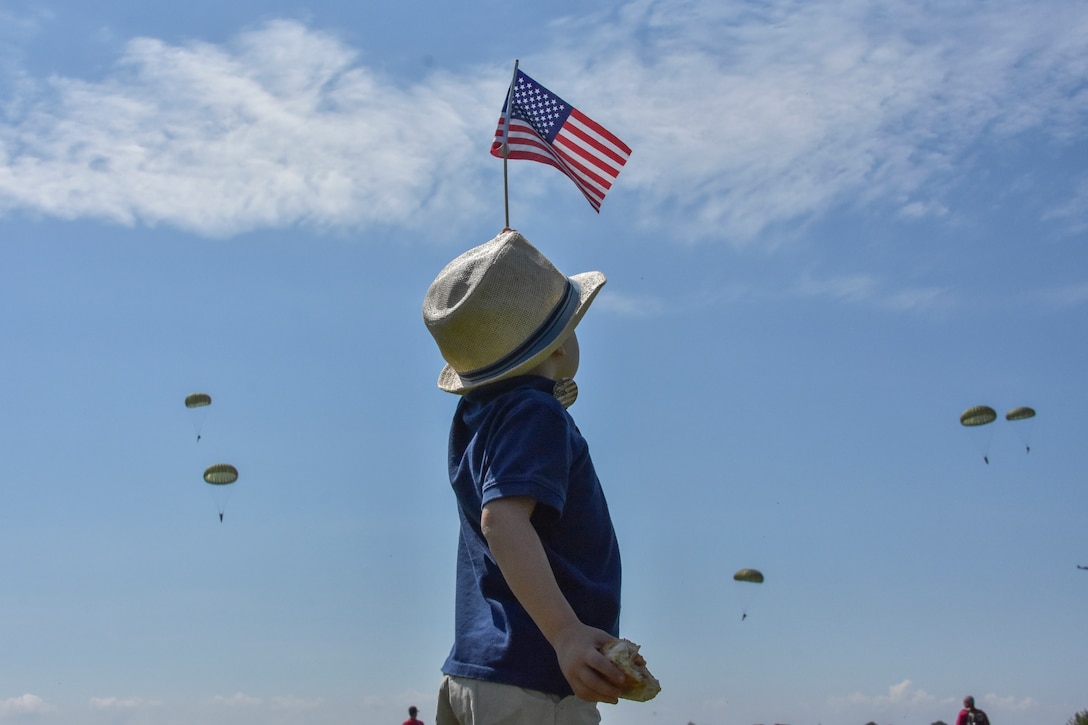 A young boy dressed in a fedora and waving an American flag welcomes paratroopers as they jump into Sainte-Mere-Eglise, France.