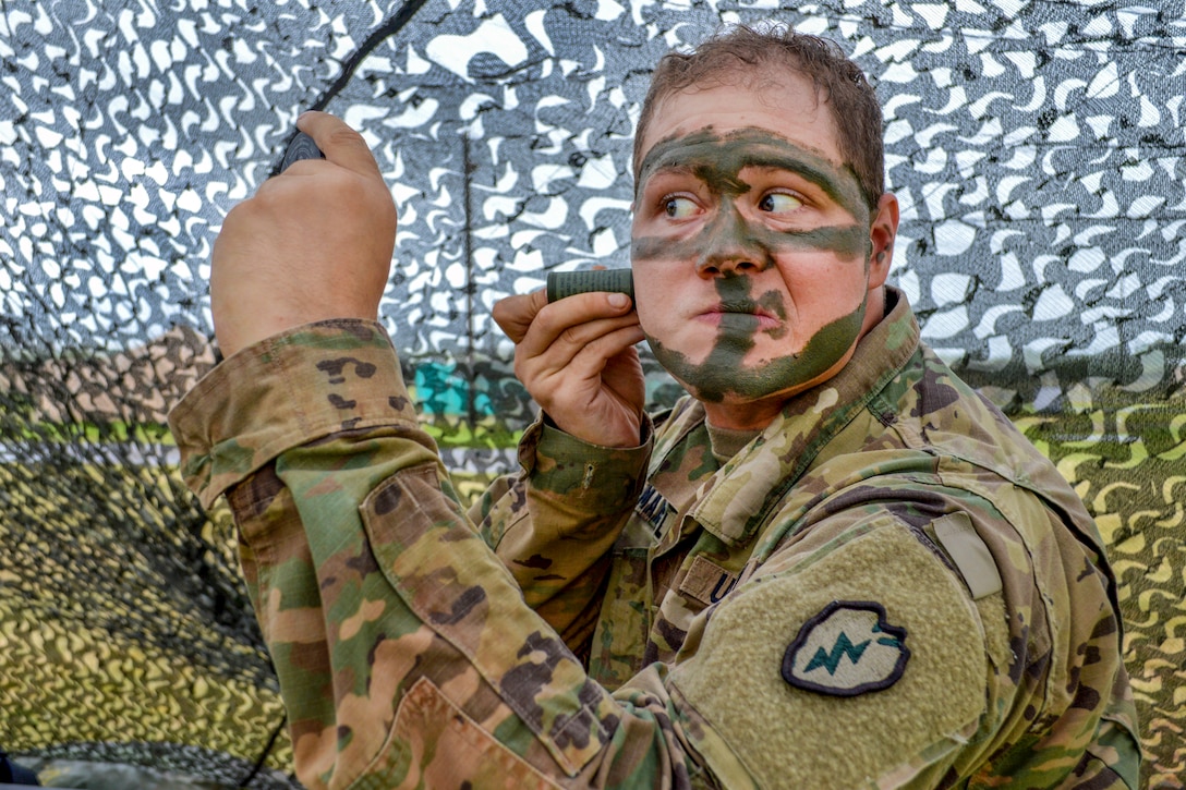 A soldier looks with paint stripes on his face looks in the mirror while applying more, against a backdrop of netting.
