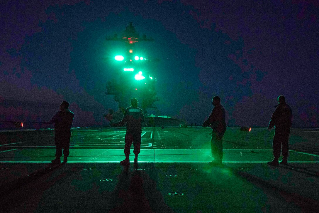 Four sailors stand in a line holding a line on a ship's deck illuminated by green light at night.