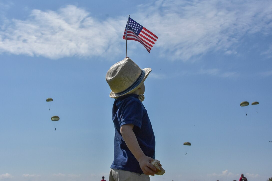 A young boy dressed in a fedora and waving an American flag welcomes paratroopers as they jump into Sainte-Mere-Eglise, France.