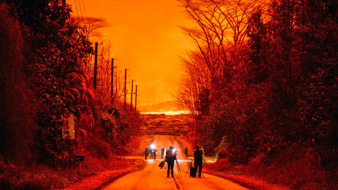 People with lights and equipment walk down a street against the backdrop of a fiery orange sky.