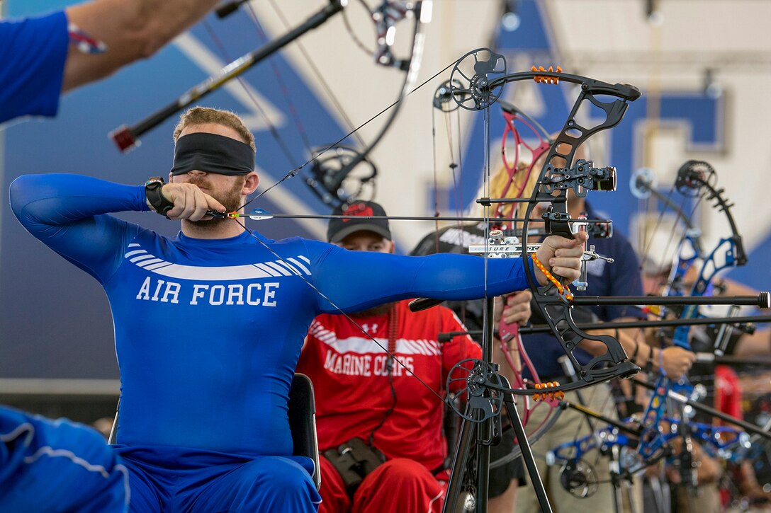A sitting, blindfolded athlete aims a bow, as others do the same behind him.