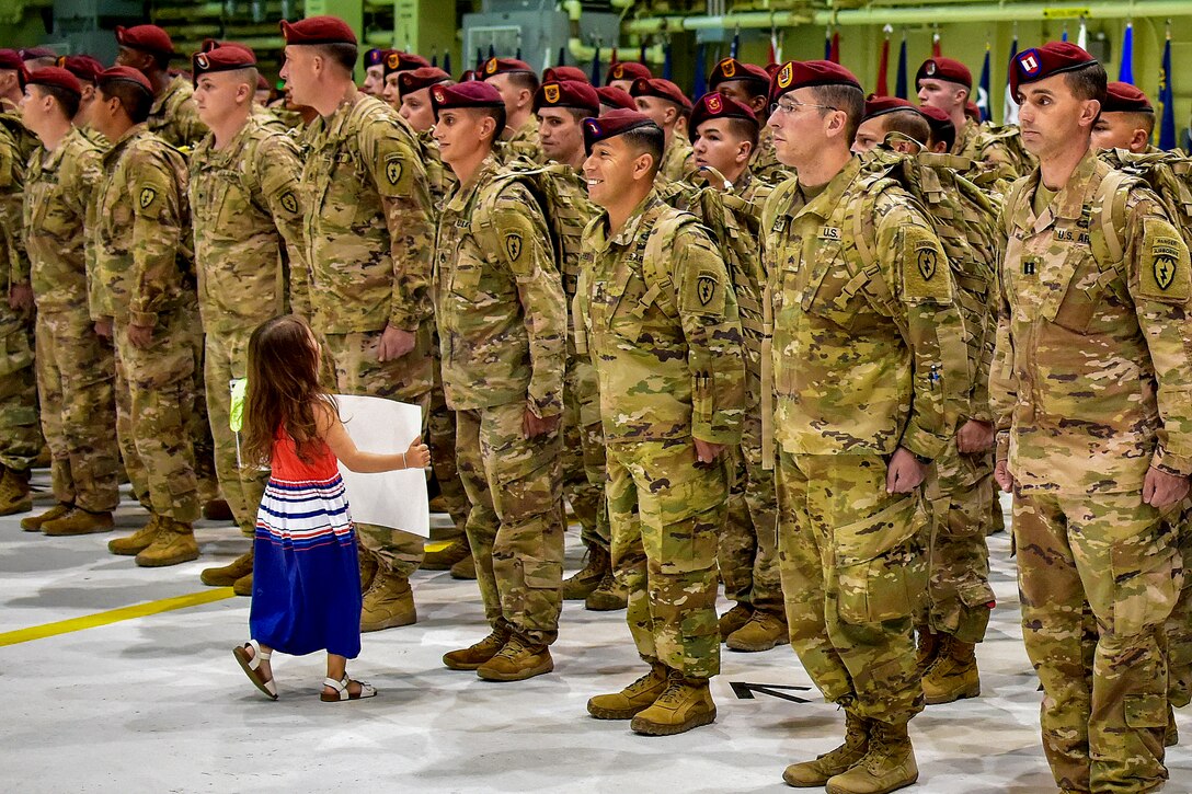 A girl carrying a sign approaches a line of soldiers in formation in a hangar.