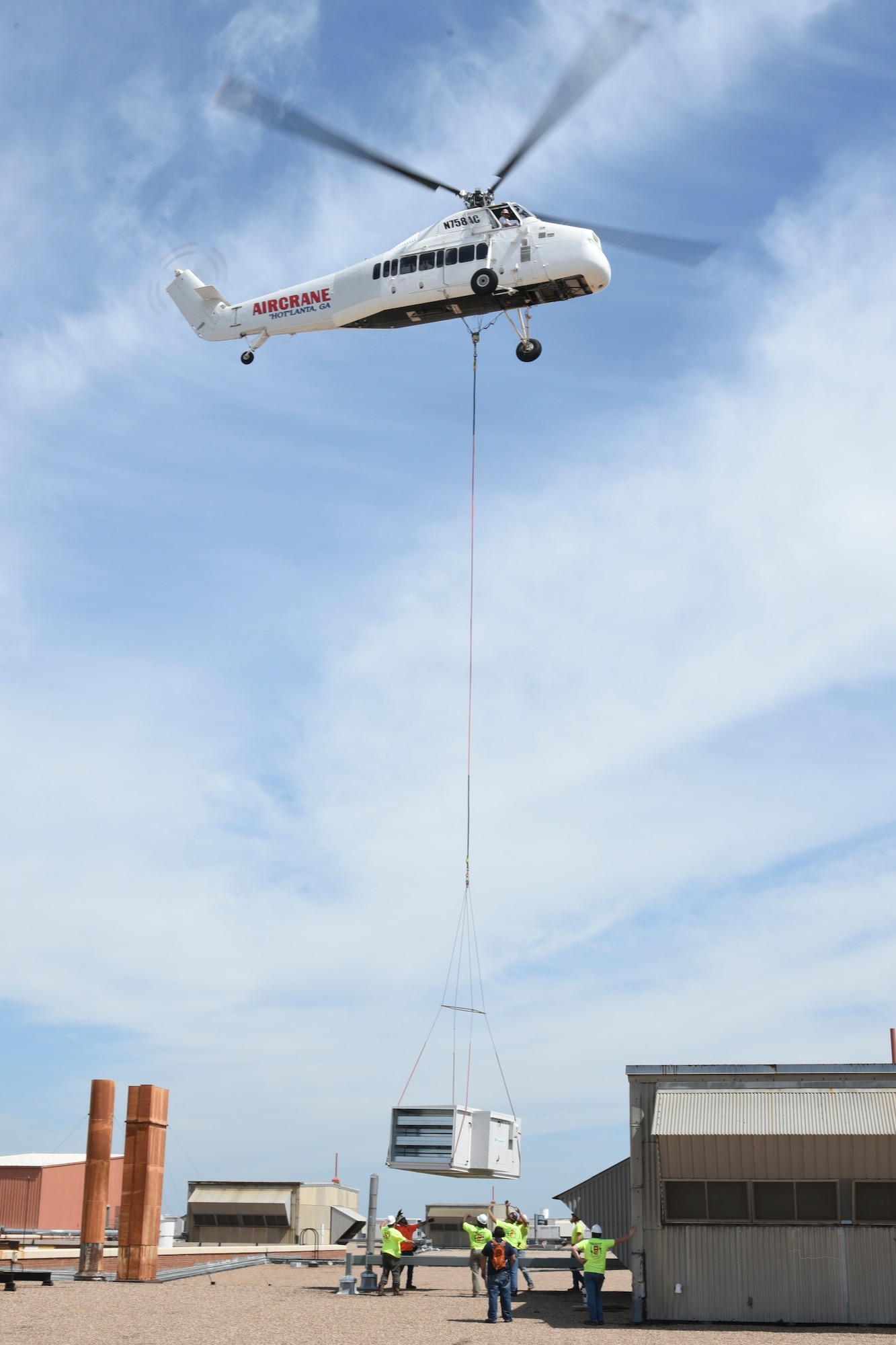Tradesmen from Matherly Mechanical contractors look up as a large heating unit is lowered in to place by a Sikorsky S-58T heavy-lift helicopter from Aircrane Incorporated May 27, 2018, Tinker Air Force Base, Oklahoma.