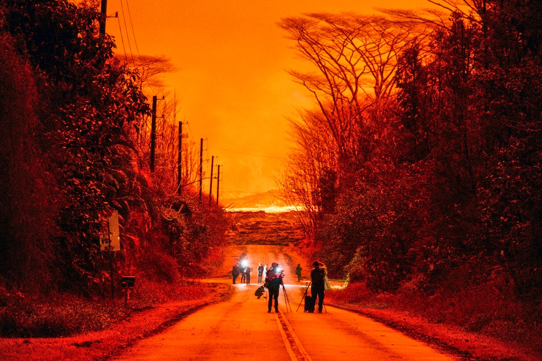 People with lights and equipment walk down a street against the backdrop of a fiery orange sky.