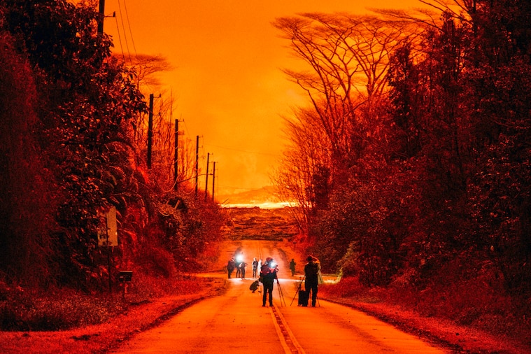 People with lights and equipment walk down a street against the backdrop of a fiery orange sky.