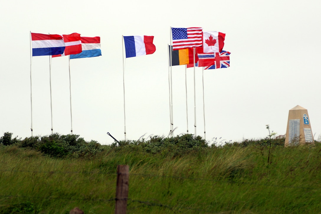 The United States of America flag, center right, along with flags from eight other nations, wave in the wind above a monument.