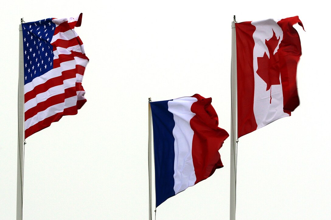 The flags of the United States, France and Canada wave in the wind while wreaths are laid at the base of a monument.