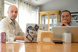 Two people sit at a home kitchen table with laptops.