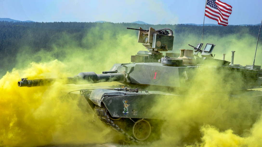 A tank displaying an American flag drives through yellow smoke.