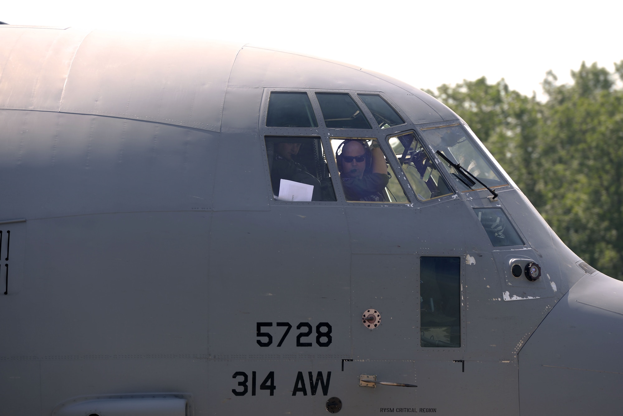 A man wearing sunglasses looks out the front of the windows in a C130J.