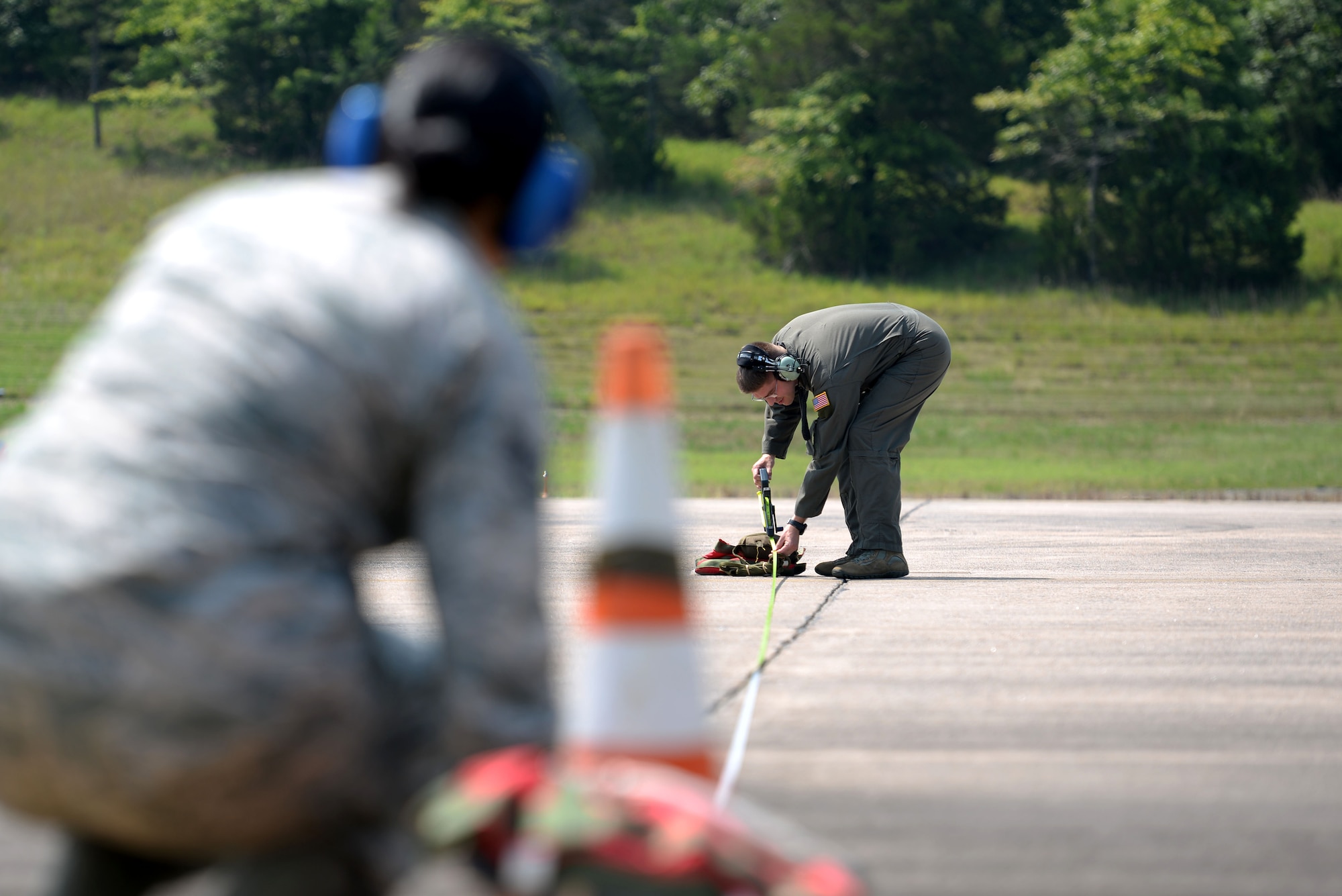 A woman wearing the Airman Battle Uniform with blue ear covers holding down a measuring tape at an orange cone while a man wearing a green flight suit pulls the other end of the measuring tape to a sand bag.