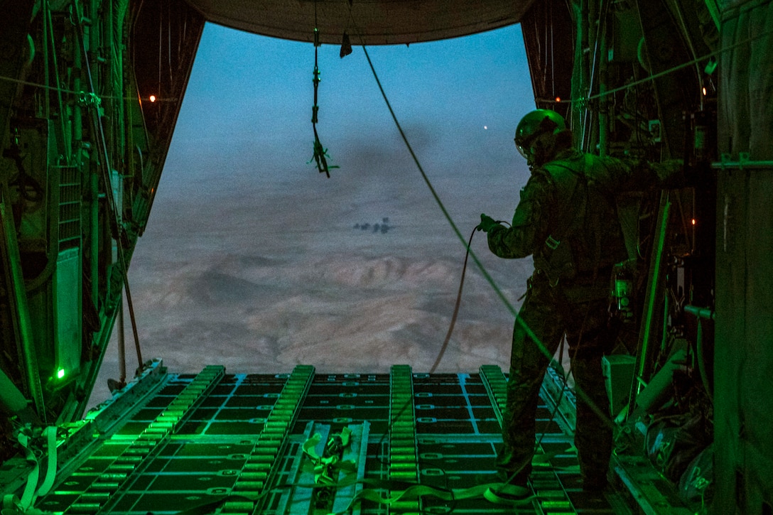 An airman looks out over desert terrain from an open aircraft door.