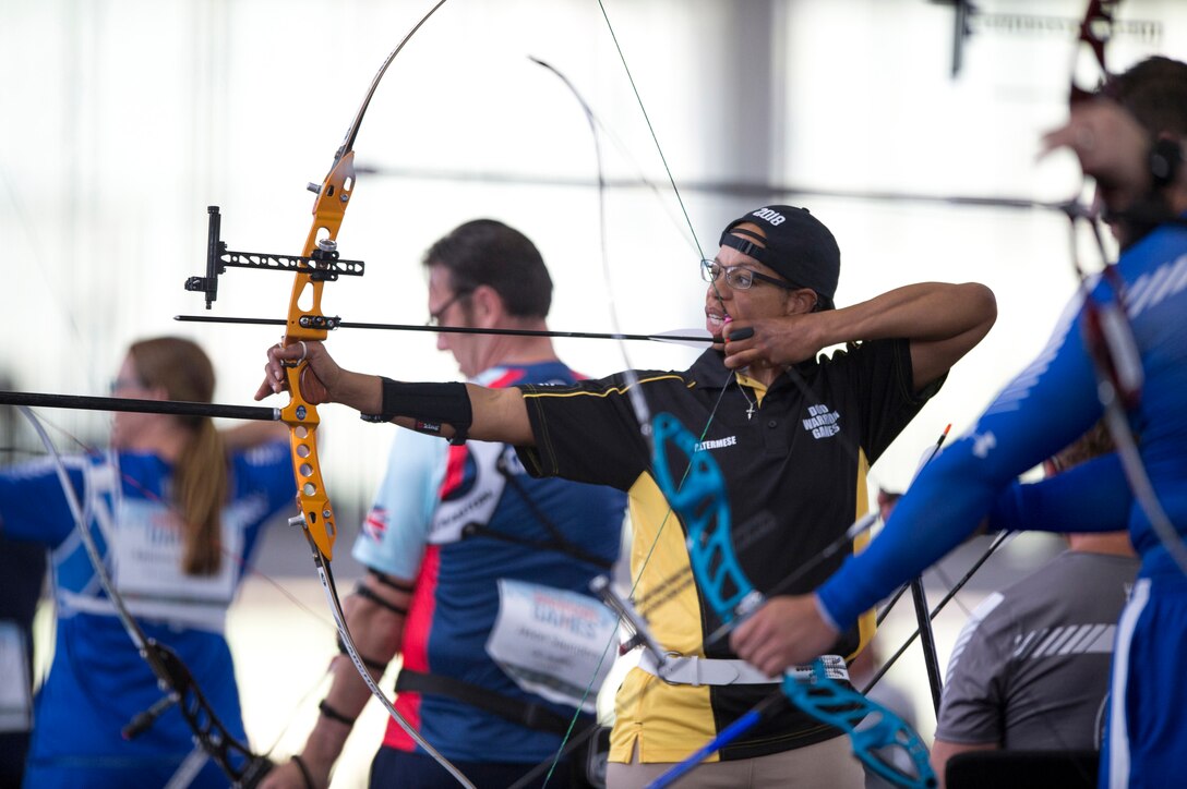 A soldier pulls an arrow back in her bow while standing in a row with fellow archers.