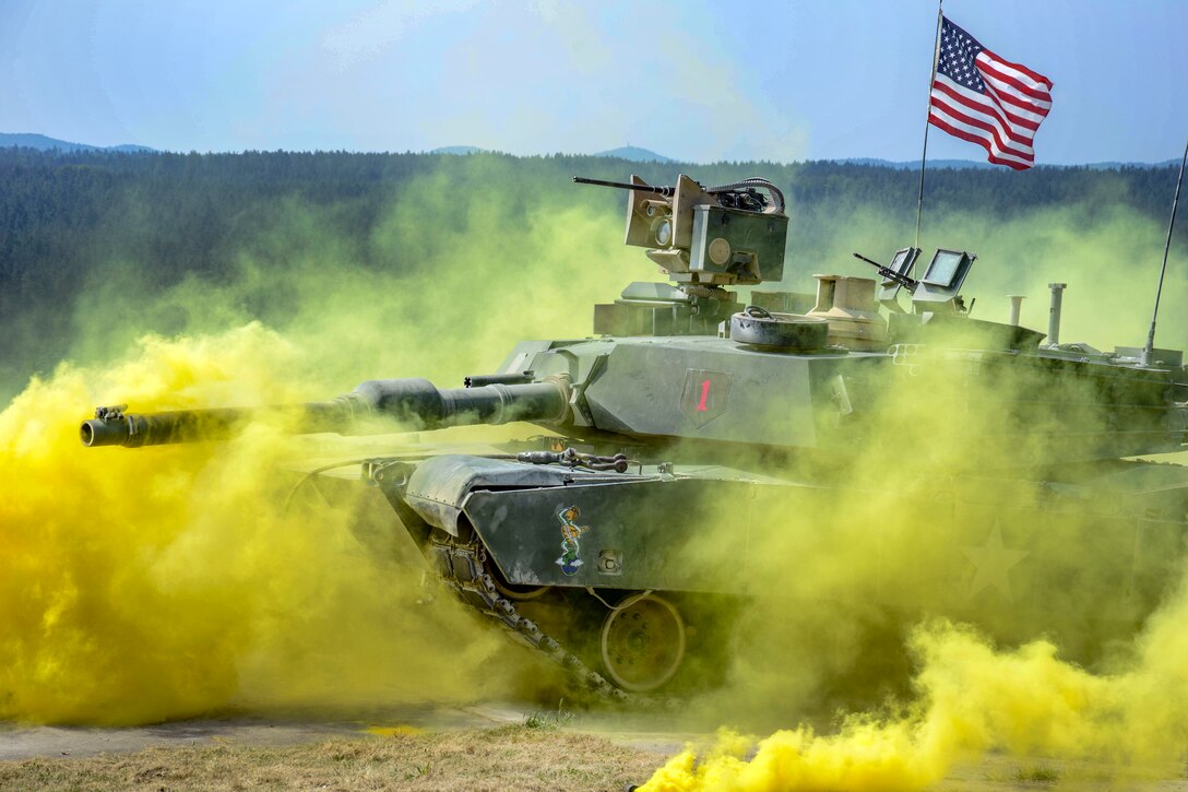 A tank displaying an American flag drives through yellow smoke.